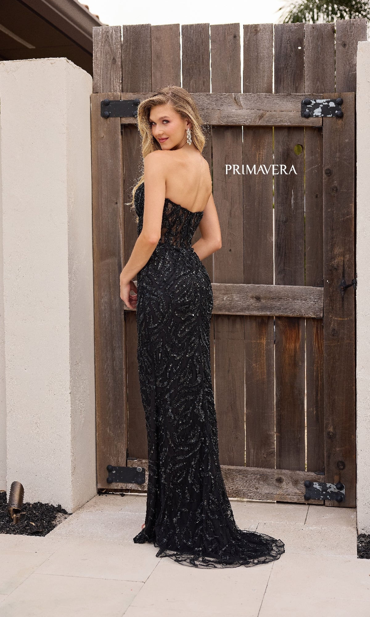 A woman wears the Primavera 4360 long strapless sequin prom dress, standing outdoors in front of a wooden gate labeled